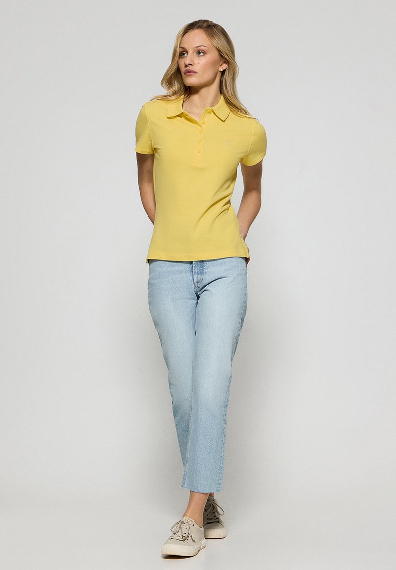 Żółta koszulka polo, krótki rękaw, dopasowany krój, jasnoniebieskie dżinsy i beżowe trampki. Prosty kołnierzyk i zapięcie na guziki na koszulce.