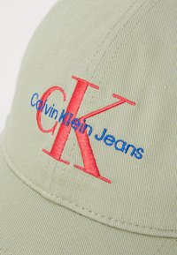 Bež kapa s zakrivljenim vizirom. Ima izvezeni crveni "CK" i plavu natpis "Calvin Klein Jeans". Tekstura tkanine s detaljima šivanja.