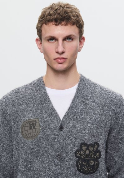 Grijze gebreide cardigan met V-hals, met twee patches: een globe-logo en een fantasievolle gezicht met bloemen. De knopen zijn zwart en de textuur is zacht.