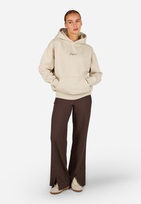 Μπεζ φούτερ με μπροστινή τσέπη και λογότυπο, συνδυασμένο με καφέ παντελόνι με χωριστό ποδαράκι και μπεζ παπούτσια, με άνετη εφαρμογή και μαλακή υφή.