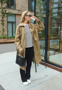 Beige trenčkot cez námořnícku a bielu pruhovanú blúzku, v kombinácii s čiernymi nohavicami a bielymi teniskami. Drží kávu v pohári a čiernu tašku.