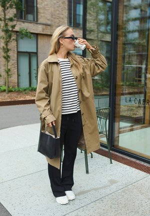 Beež trench coat sinine ja valge triibuline topp, koos mustade pükste ja valgete tossudega. Hoides kohvitassi ja musta kotti.