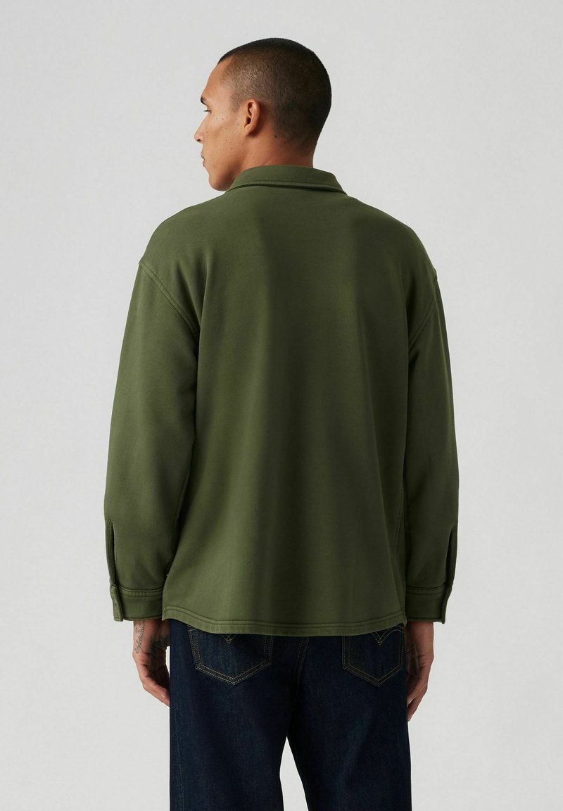 Maglione verde con collo, vestibilità comoda, maniche lunghe e spacchi laterali. Indossato su jeans scuri, con una texture liscia.