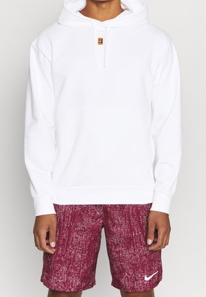 Homme portant un sweat à capuche blanc uni avec un petit logo sur la poitrine et un short Nike rouge à motifs avec un logo swoosh blanc.