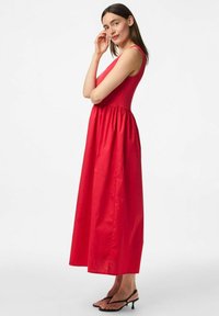 Sarkans maksi kleita ar pieguļošu augšdaļu un plūstošu svārku, izgatavota no gludas auduma. Tam ir plānas siksnas un savilkta jostasvieta, apvienota ar melniem sandaliem.