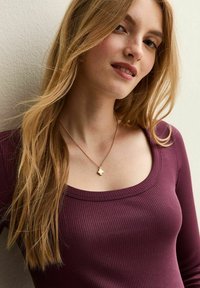 Czerwony ribbed top z długim rękawem i szerokim dekoltem, połączony z delikatnym złotym naszyjnikiem z teksturowanym wisiorkiem. Długie, falowane włosy.