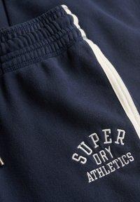 Mørkeblå træningsshorts med hvide sidelister og broderet "SUPER DRY ATHLETICS." Har en elastisk talje og blød tekstur.