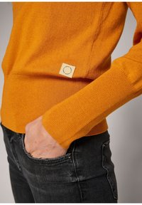 Orange ribbed sweater with long sleeves, featuring a square woven label at the hem. Paired with fitted dark jeans.