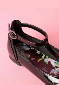 Zapato Mary Jane de charol negro con un diseño de ondas, tira ajustable con herrajes plateados y plantilla con patrón floral.