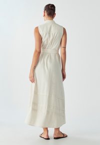 Biela, bezrukávová maxi šaty s golierovým výstrihom, opasok v páse, viacvrstvový dizajn a ľahký materiál, nosené s čiernymi šľapkami.