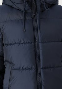 Mornarska puffer jakna z zadrgo na sprednji strani, nastavljivim kapuco s šnurovanjem in kvadratnim vzorcem. Ima stranska žepa in lahkoten material.