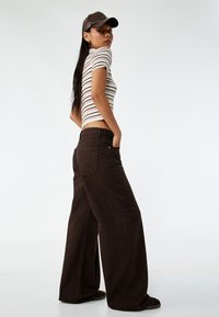 Stribet ribbet crop top med korte ærmer, parret med vidde mørkebrune bukser. Modellen bærer en brun cap, der fremhæver en glat tekstur og pasform.