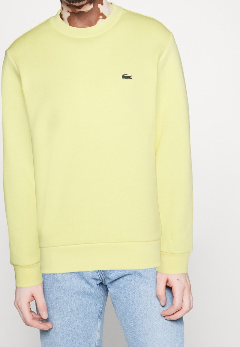 Ljusgul sweatshirt i mjukt tyg har rund halsringning, ribbade manschetter och avslut, samt en liten Lacoste-logotyp på bröstet.