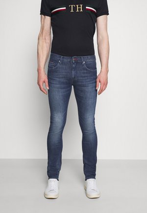 Jeans ajustados de un oscuro azul con textura desgastada, cinco bolsillos y cierre de botón. Combinados con una camiseta negra con rayas verdes, rojas y blancas.