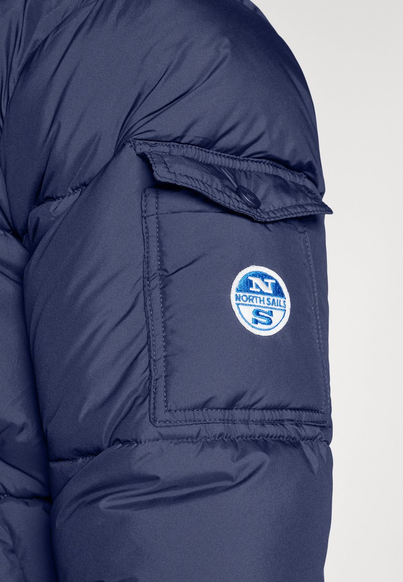 Ärmel einer marineblauen Steppjacke mit einer Klappentasche, die ein kreisförmiges "North Sails"-Logo in blauer und weißer Stickerei zeigt. Glatte Textur, gestepptes Design.