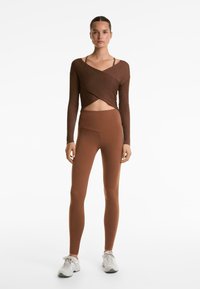 Smeđa rebrasta majica dugih rukava s preklopnim dizajnom, u kombinaciji s smešnim tajicama visoko strukiranim. Bijele sportske tenisice upotpunjuju cijeli outfit.