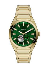 Guldtonet armbåndsur med en grøn tekstureret skive, lysende time-markører og en delvis udsigt til bevægelsen. Udstyret med et metalled armbånd.