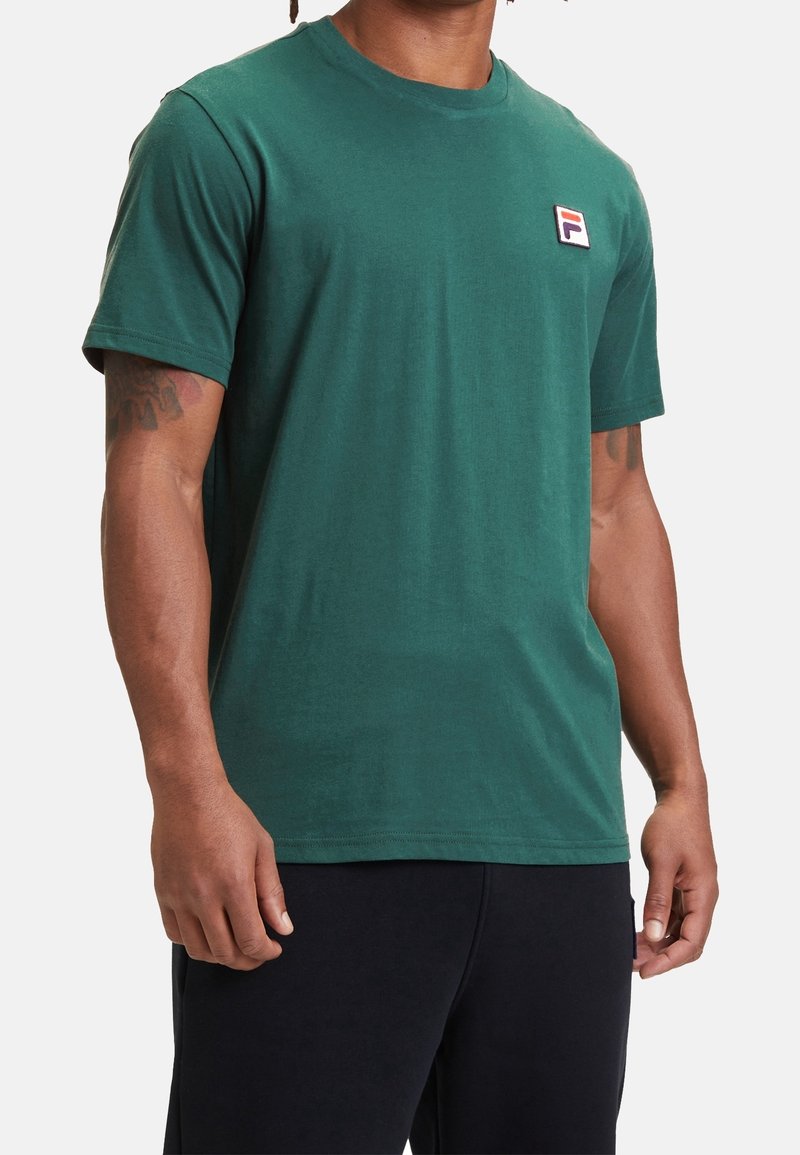 T-shirt en coton vert à manches courtes, col rond, avec un petit écusson logo sur la poitrine gauche. Texture lisse, coupe décontractée.