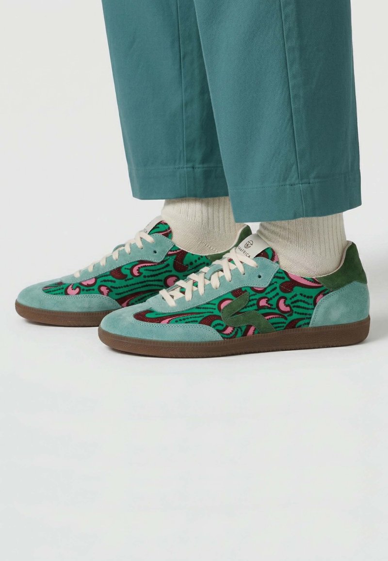 Baskets à motifs bleu sarcelle et vert avec lacets blancs et semelles marron, portées avec des chaussettes beiges et un pantalon court bleu sarcelle sur fond blanc.