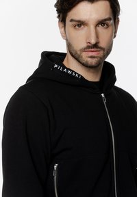 Czarna bluza z kapturem na zamek błyskawiczny, z kieszenią kangurką, widocznym detalem zamka oraz metką z napisem "PILAWSKI" na kapturze. Gładka tekstura.