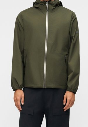 Homme portant une veste à capuche vert olive avec fermeture éclair à l'avant et un pantalon noir avec poches, debout devant un fond blanc.