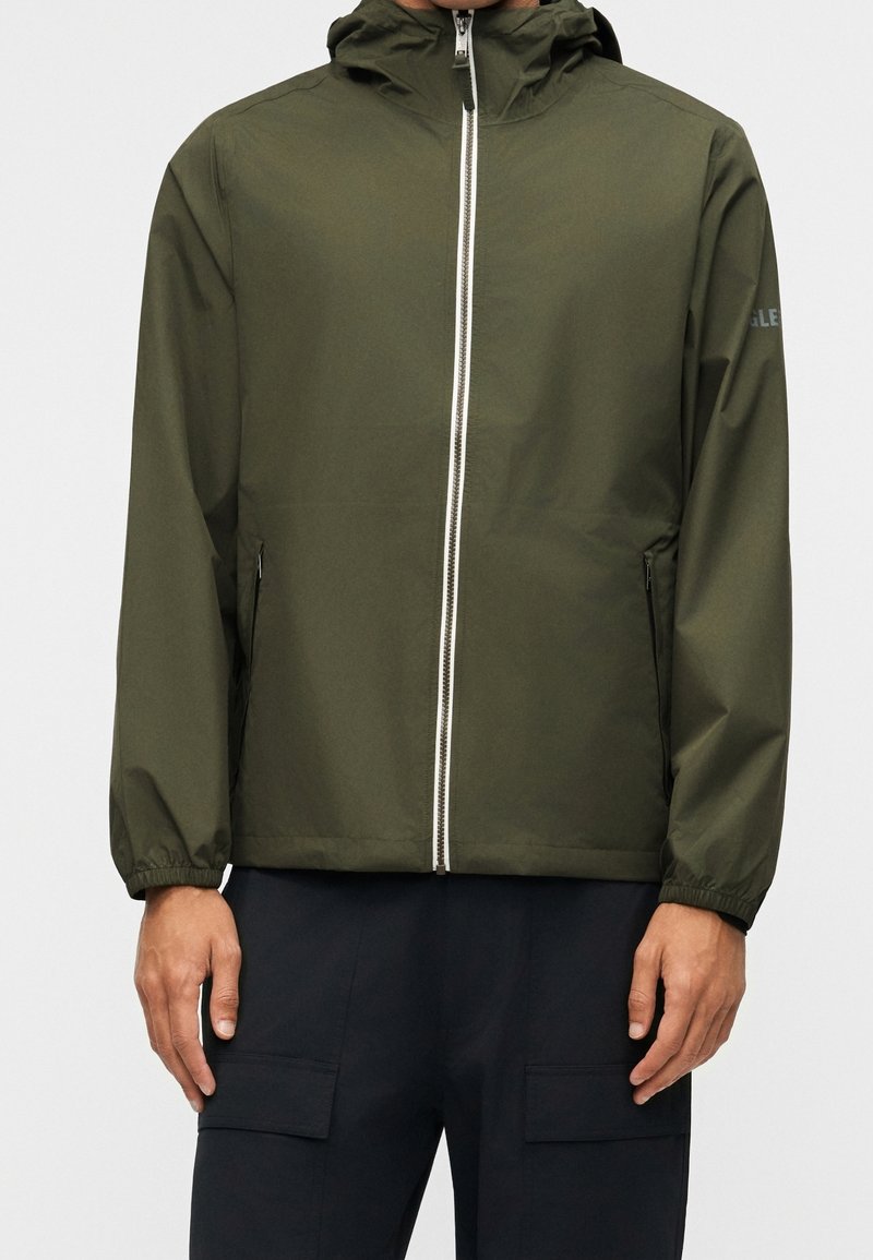 Homme portant une veste à capuche vert olive avec fermeture éclair à l'avant et un pantalon noir avec poches, debout devant un fond blanc.