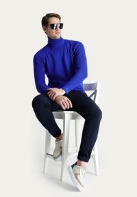 Maglione a collo alto blu a righe, pantaloni neri slim-fit e sneaker bianche con accenti beige e verdi. La persona indossa occhiali da sole scuri, seduta su uno sgabello.