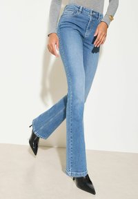 Hellblaue Schlagjeans mit hoher Taille, aus Denim gefertigt, mit rohem Saum und dezentem Verblassen, kombiniert mit schwarzen spitzen Absatzschuhen.