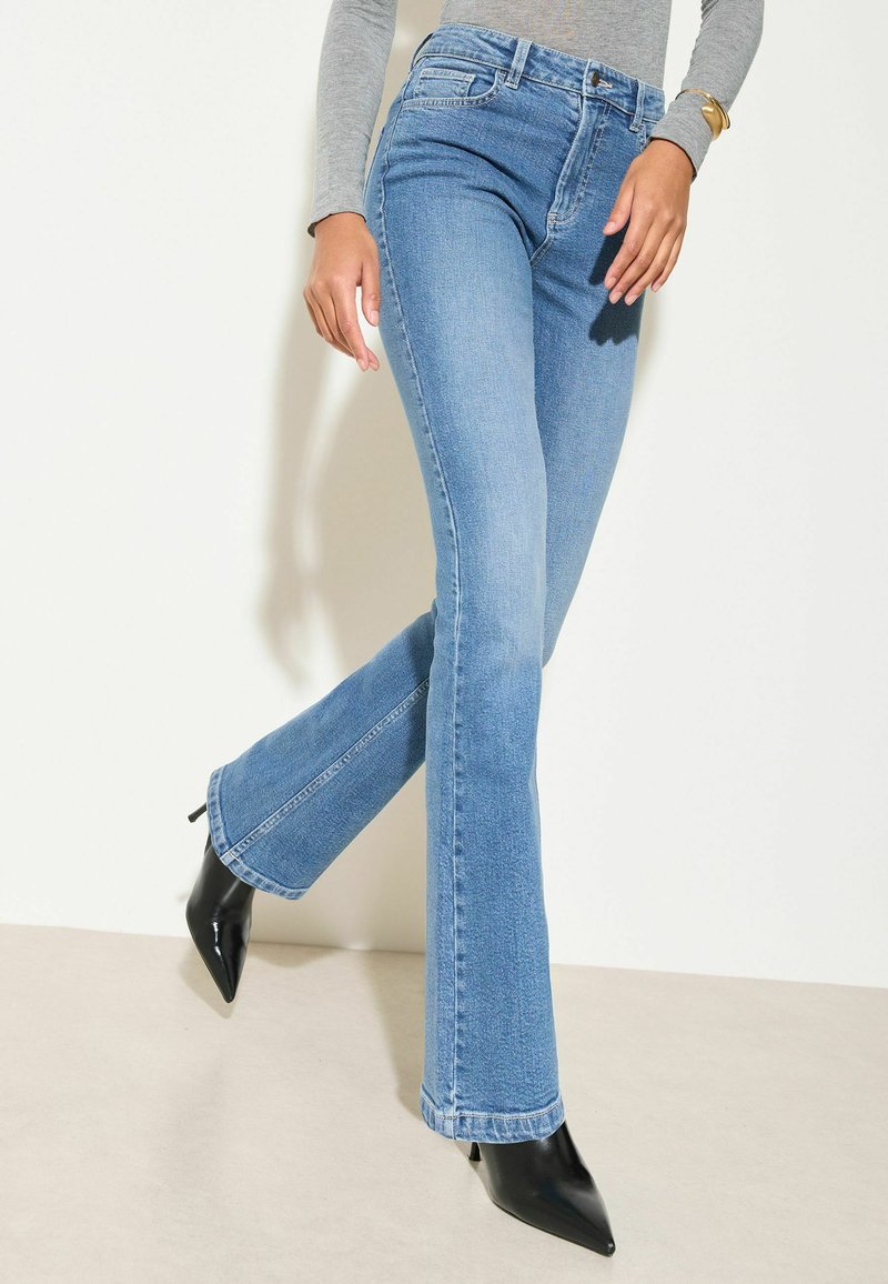 Hellblaue Schlagjeans mit hoher Taille, aus Denim gefertigt, mit rohem Saum und dezentem Verblassen, kombiniert mit schwarzen spitzen Absatzschuhen.