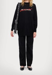 Czarny, oversize sweter z graficznym napisem, zestawiony z czarnymi, luźnymi spodniami i płaskimi butami. Dopełniony czarną torbą na pasku z srebrnymi elementami.