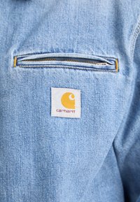 Svetlomodrá denimová bunda s prehĺbeným vreckom na zips a štvorecovým znakem Carhartt v bielej farbe s žltým dizajnom.