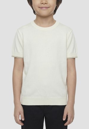 Enfant aux cheveux bruns courts portant un t-shirt blanc uni à manches courtes et un pantalon noir, souriant légèrement, sur un fond uni.