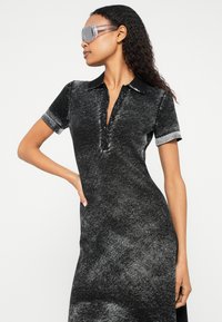 Femeie cu păr lung și creț, purtând ochelari de protecție transparenți și o rochie polo neagră cu efect acid-wash, cu mâneci scurte și nasturi.