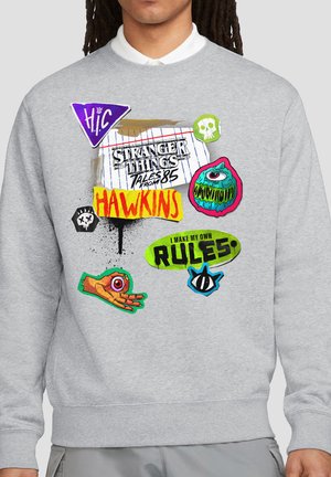 Gray sweatshirt with colorful "Stranger Things Tales from '85" text, Hawkins name, skulls, an eye monster, disembodied eye hand, and "I make my own rules" badge.