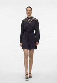 Sieviete mugurā melnu kleitu ar mežģīņu ielaidumu, garām piedurknēm, akcentētu jostu un melnām potīšu siksniņām, stāvot pret vienkārša balta fona.