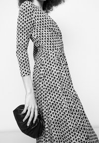 Schwarz-weiß gemustertes Kleid mit langen Ärmeln, taillierter Oberseite und fließendem Rock. Das Model hält eine schwarze, geraffte Clutch-Tasche.