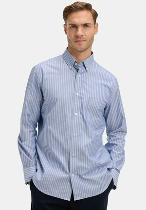 Homme portant une chemise à boutons bleu clair à rayures verticales avec col, debout les mains dans les poches devant un fond uni.