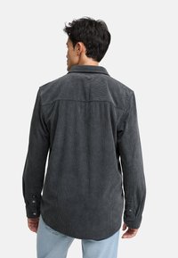 Camisa de pana en gris oscuro con cuello abotonado, textura de estrías verticales visibles y puños abotonados. La vista trasera muestra un diseño de corte relajado.
