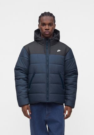 Ung mand iført en sort og mørkeblå Nike dunjakke med hætte, stående mod en ensfarvet lys baggrund.