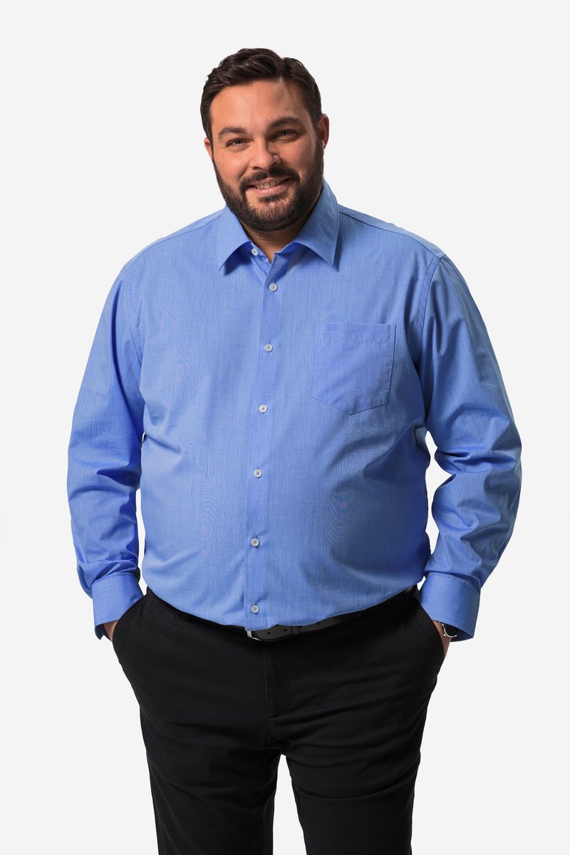 Hombre sonriente con cabello corto y oscuro y barba, que lleva una camisa azul de manga larga abotonada y pantalones negros con las manos en los bolsillos.