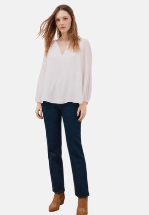 Blouse blanche plissée avec un col en V et des manches longues, associée à un jean foncé et des bottines brunes. Design simple avec une texture lisse.
