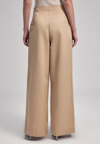 Pantalons à jambe large en beige clair, fabriqués dans un tissu lisse, avec une taille haute et des coutures nettes, présentant une finition légèrement brillante.