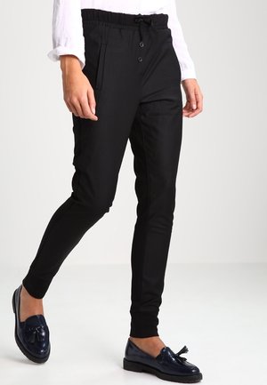 Personne portant un pantalon noir fuselé avec taille à bouton et cordon, associé à des mocassins foncés à pampilles et une chemise blanche à manches longues.