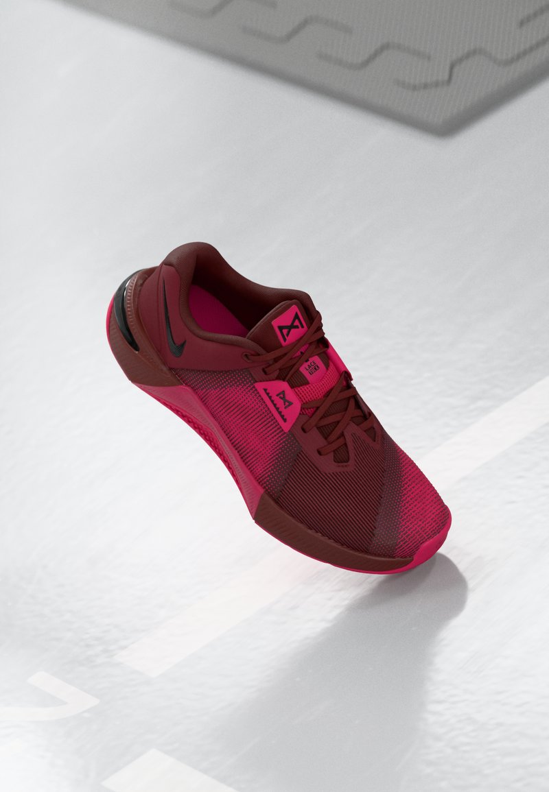 Red and pink athletic shoe with mesh upper, textured design, black accents, and reinforced heel. Features traditional lace-up closure.