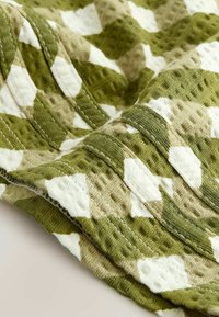 Tissu texturé vert et beige avec motifs géométriques blancs, coutures visibles et détail de bande bouclée.