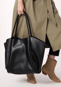 Grote zwarte leren tote bag met twee lange hengsels, met een gladde textuur en subtiele branding. Draagt met een beige trenchcoat en tan laarzen.