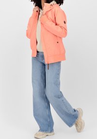 Letvægtig koral bomberjakke med hætte, lynlåslukning og ribbede manchetter. Sammen med løse lyseblå jeans og beige sneakers.