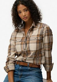 Camisa de franela a cuadros en tonos marrón y beige con cuello abotonado, que cuenta con dos bolsillos en el pecho y mangas remangadas, usada sobre vaqueros azules.