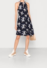 Vestido floral azul marino con escote halter, que presenta un corte en forma de llave y una falda fluida. Combinado con zapatillas beige y un bolso a juego.