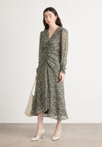 Robe midi à imprimé floral en noir et crème avec des détails froncés, des manches longues, un col en V et un ourlet fluide. Associée à des talons beiges et un sac fourre-tout.